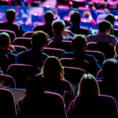 Backs of an audience in a dark conference room facing the stage.